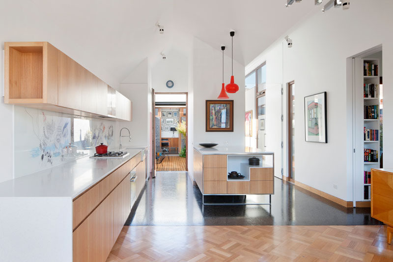 At the back of this modern house is the kitchen, with light wood kitchen cabinets and island, while an artistic backsplash and a couple of red accents bring a touch of fun and color into the room. From the kitchen you can also see out onto the deck just outside and into the home office on the other side of it.