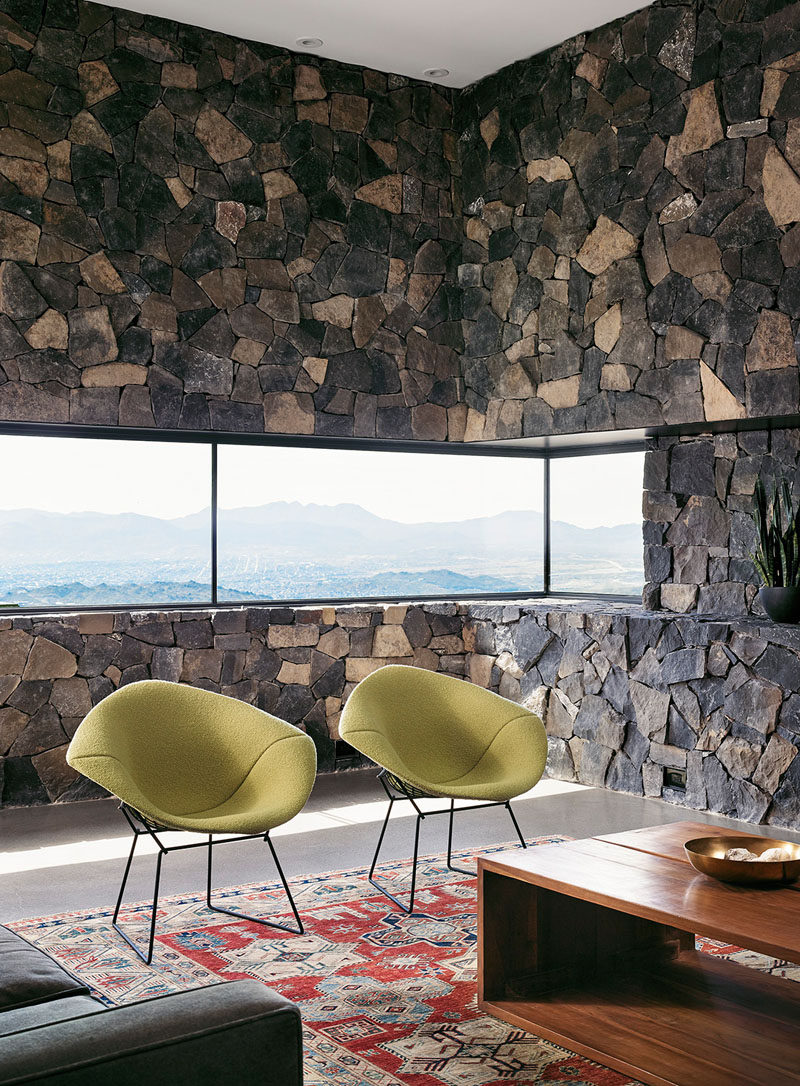 In the living room of this modern house, stone covers the walls and creates a rustic, mountain feel. A letter box window that wraps around the corner looks out to the valley and city below. Modern green chairs and a rectangular wood coffee table warm up the space and make it an inviting spot sit and watch the sunset. 