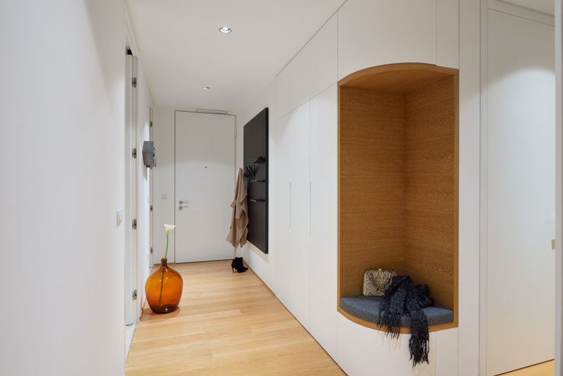 This entryway in a modern apartment has a small section of cabinets, however instead of the cabinets wrapping around the corner, the designers removed a section included a small seat with a cushion surrounded by wood, making it the ideal place to put your shoes on.