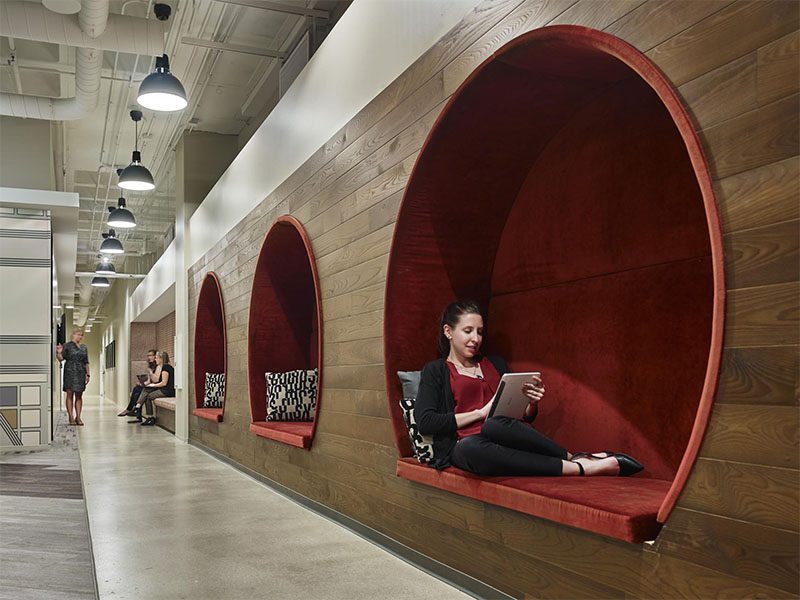 In this modern office, three circular seating nooks with red upholstered cushions are embedded within a dark wood wall, creating a cozy and intimate environment to work or take time away from a screen.