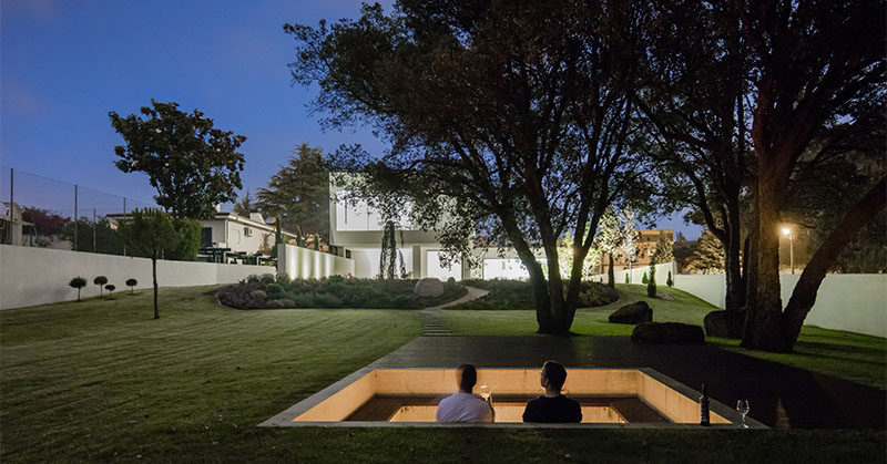 Sunken down into the grassy landscaped yard of this modern house, is a square lounging pit with built-in seating that wraps around the fire and allows people to lean against the wall. From the lounge, you are able to have a view of the house, and at night it's lit up, highlighting the design of the house. #FirePit #Landscaping #Backyard #OutdoorLounge #SunkenFirePit