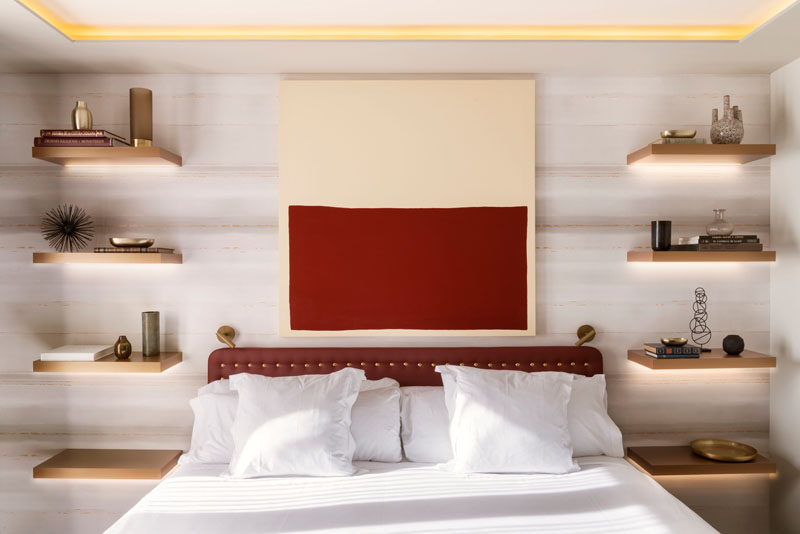 In this modern bedroom, four floating wood shelves with hidden lighting have been installed, with the lowest shelf at the correct height for a bedside table, while the other three shelves have been used to display decorative items. #BedroomDesign #BedsideTable #InteriorDesign #Shelving