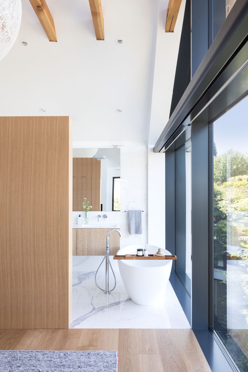 In this modern ensuite bathroom, a freestanding white bathtub is positioned in front of the windows to take advantage of the views, while further behind the wall is the vanity area, toilet and a walk-in shower. #Bathroom #ModernBathroom #FreestandingBathtub