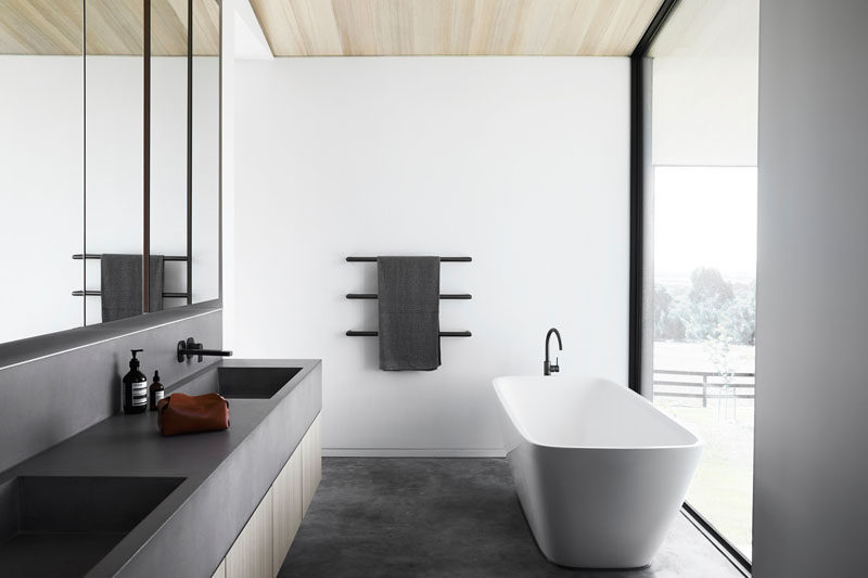 This modern bathroom has a dark grey vanity that features a built-in sink, and a white freestanding bathtub, that's positioned in front of the large floor-to-ceiling window. #Modernbathroom #BathroomDesign