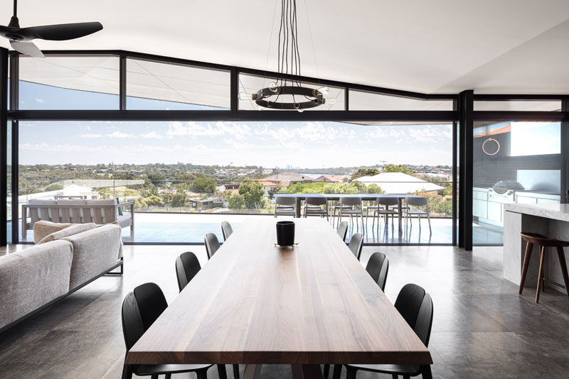 The balcony provides expansive views from the elevated position of this modern house, and enough room to have an outdoor lounge and dining area, as well as an outdoor kitchen with a BBQ. Inside, the large wood dining table separates the living room from the kitchen. #ModernInterior #DiningRoom #Balcony #OutdoorKitchen