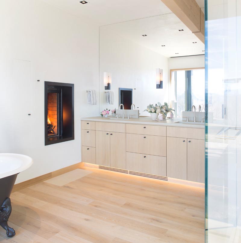 In this modern en-suite master bathroom, a wood vanity sits below a full wall mirror that reflects the light from the window, while the fireplace adds warmth and a sense of relaxation to the space. #MasterBathroom #BathroomDesign #WoodVanity #Fireplace