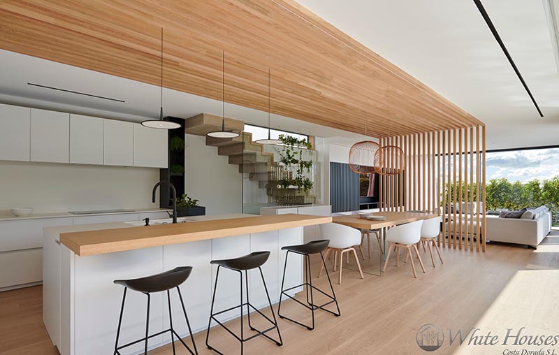 Connecting and defining this modern kitchen and dining area is a wood slat accent that travels from one wall at the end of the kitchen onto the ceiling and through to the dining area, where it becomes a partition wall, separating it from the living room. #KitchenDesign #DiningArea #WoodSlatWall #Partition #ModernKitchen