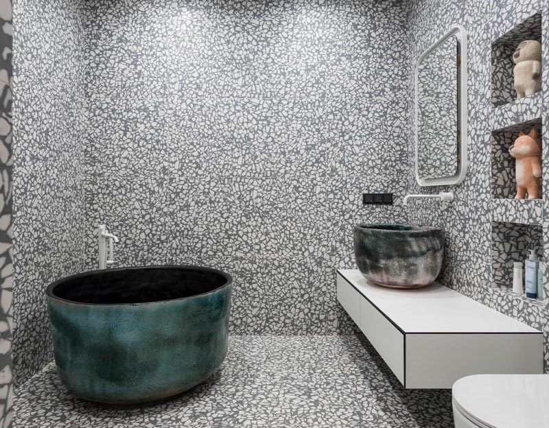 A grey and white large aggregate terrazzo tile has been installed in this modern bathroom, covering the walls and floor. #GreyAndWhiteBathroom #GreyBathroom #BathroomIdeas #SoakingTub #ShelvingNiche #ModernBathroom