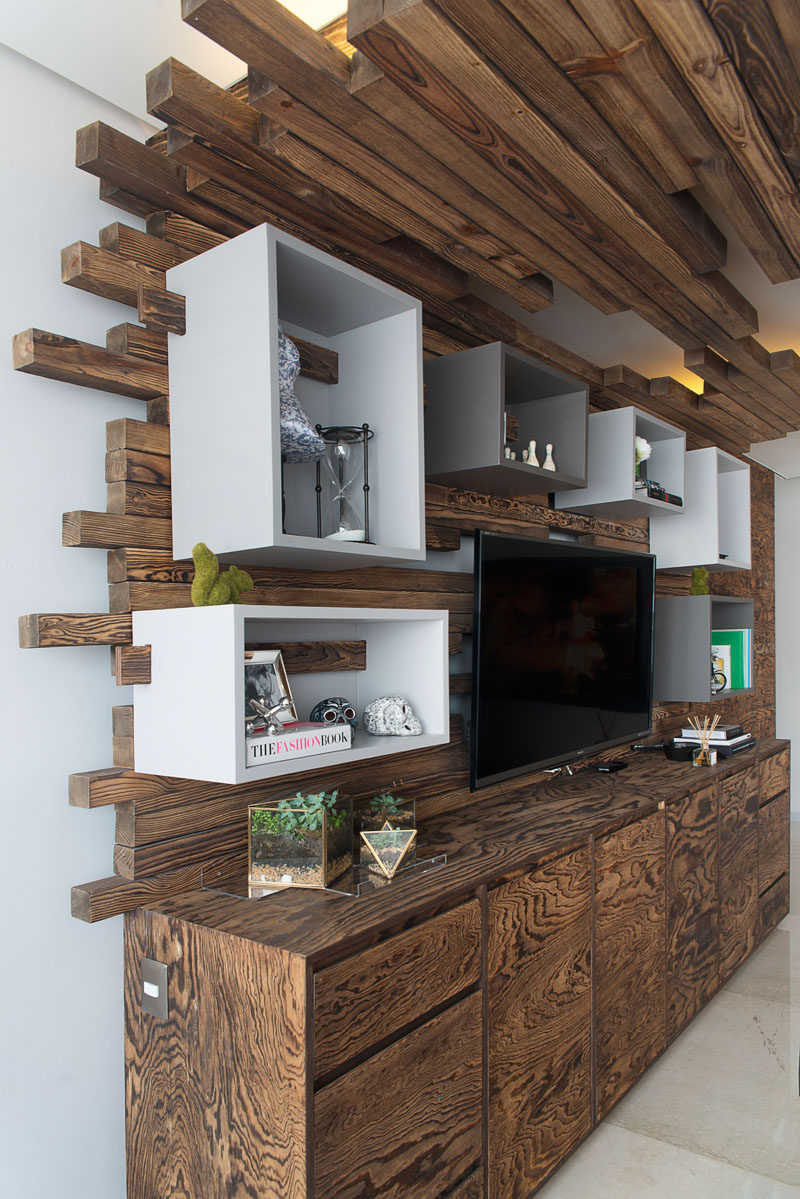 In this living room, there is a wall with a television, cabinetry and box shelving. The box shelving is attached to the wall using wood that matches the ceiling detail.