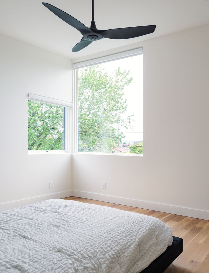 In this master bedroom, corner windows provide a view of Portland’s downtown skyline and West Hills, while a black ceiling fan and bed-frame contrast the white walls.