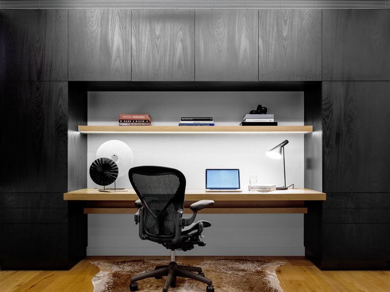 This home office is surrounded by black cabinetry, directly contrasting the wooden desk and shelf, and the white wall.