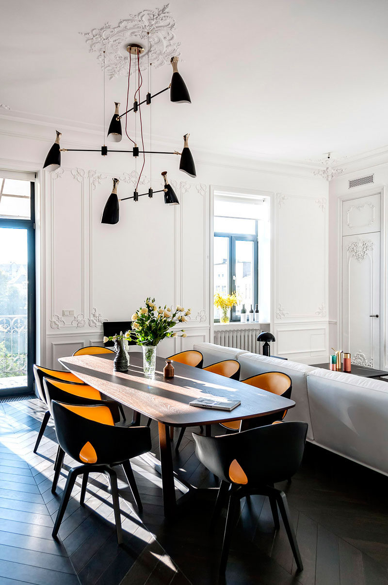 In the main living area of this apartment, the space feels large with the tall bright white walls and high ceiling. The classic wall designs contrast with the modern furniture and dark floors. A sculptural pendant light anchors the dining table in the space.