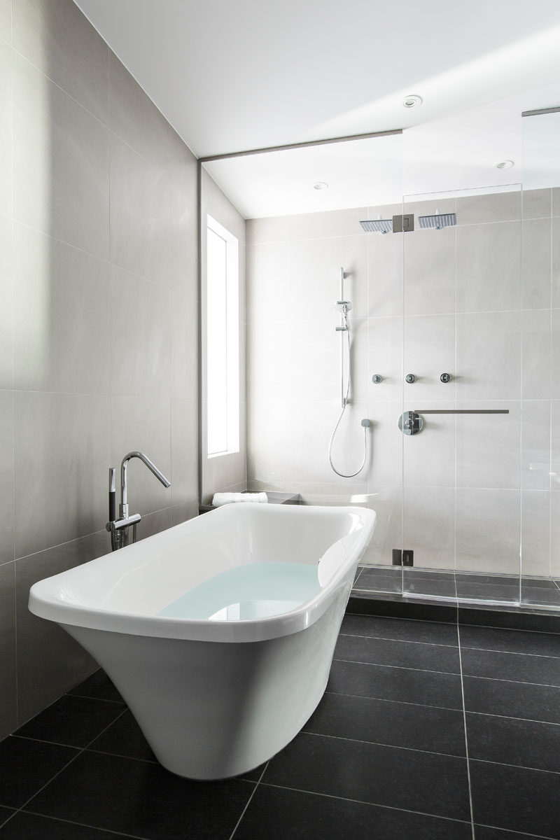 In this master bathroom, light grey tiles cover the walls, while charcoal tiles cover the floors. A glass shower surround allows the light from the window to fill the bathroom.