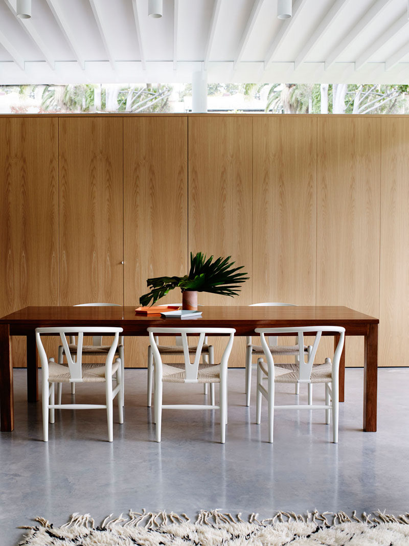 A wooden wall with hidden storage, and a dark wood dining table with white chairs make up this dining room.