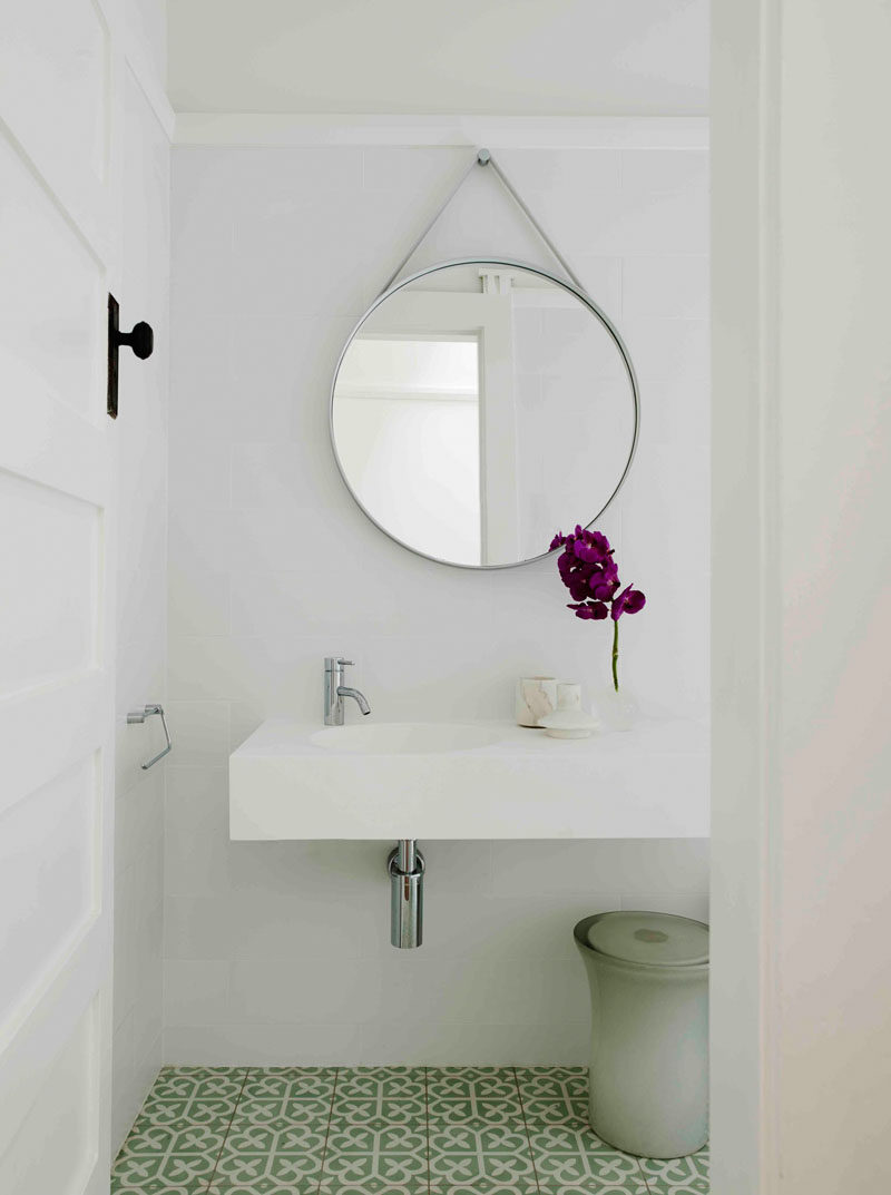 This contemporary bathroom has a round mirror hanging above the minimal white vanity, and a patterned green and white tile covers the floor to add some color to the space.