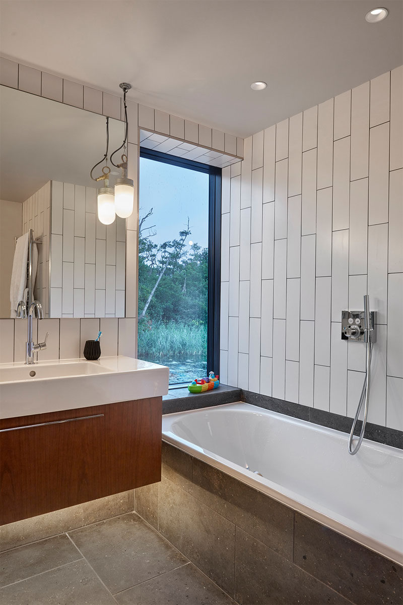 In this modern bathroom, large white tiles have been installed vertically adding to the sense of height in the room, and a tall window allows for views when taking a bath. A wood vanity is topped with a white countertop, and concrete tiles have been used for the floor and bath surround.