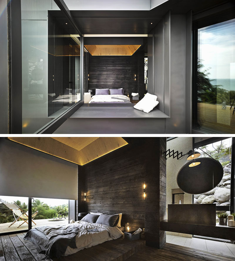 Looking into this modern bedroom through a large pivoting window, the blackened wood accent wall is a bold statement, while the lighter wood and hidden lighting in the ceiling draws your eye upwards, making the room appear larger than it is.