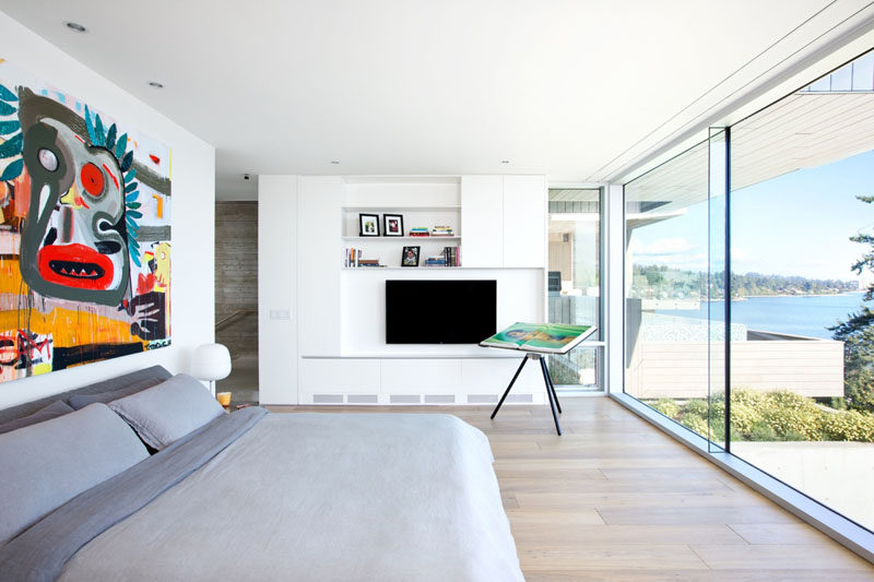 This modern bedroom has large floor-to-ceiling windows, wide plank oak flooring and built-in shelving. A large painting above the bed adds a pop-of-color to the mostly-white room.