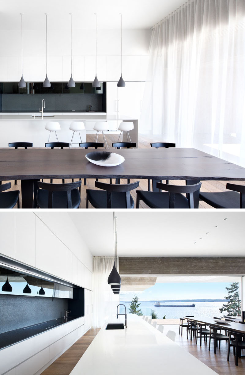 This modern dining room with a live-edge wooden table is surrounded by black dining chairs, and beside it, the kitchen has bright white cabinets, a central island lit with pendant lights and a dark grey and black backsplash.