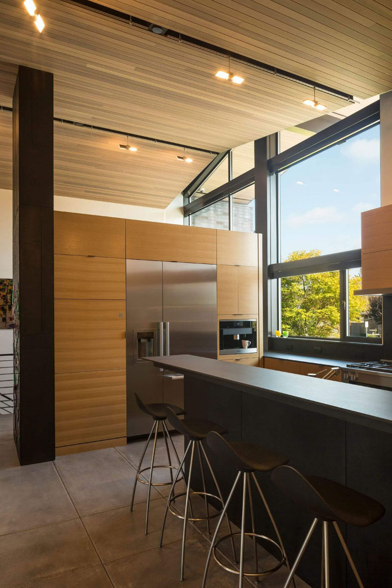 In this modern kitchen, white oak cabinets have been paired with grey porcelain counters, and a large window overlooks the front yard.