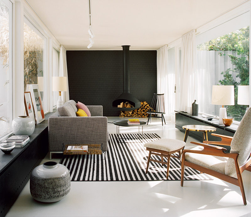 This modern living room features a hanging black fireplace, a black tile accent wall, modern furniture, light grey floors and a pale pink ceiling.