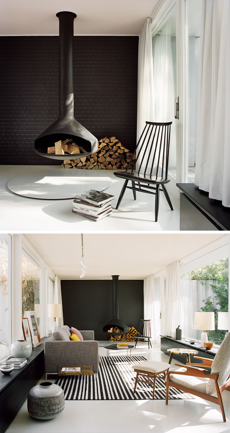 This modern living room features a hanging black fireplace, a black tile accent wall, modern furniture, light grey floors and a pale pink ceiling.