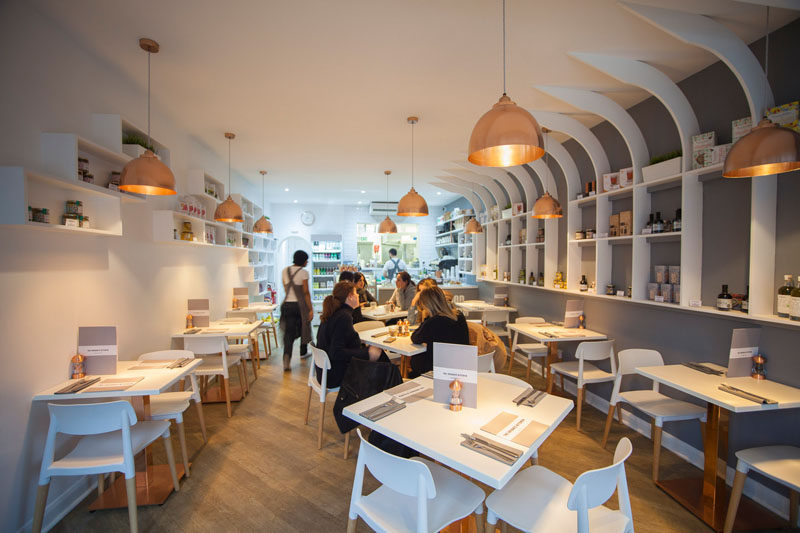 In this modern resturant interior, the designers used white for the walls, furniture, and shelves, wood on the floors and chair legs, copper accents in the form of light fixtures, table stands, and salt and pepper shakers. A grey accent wall is used to draw your eye to the white shelves.