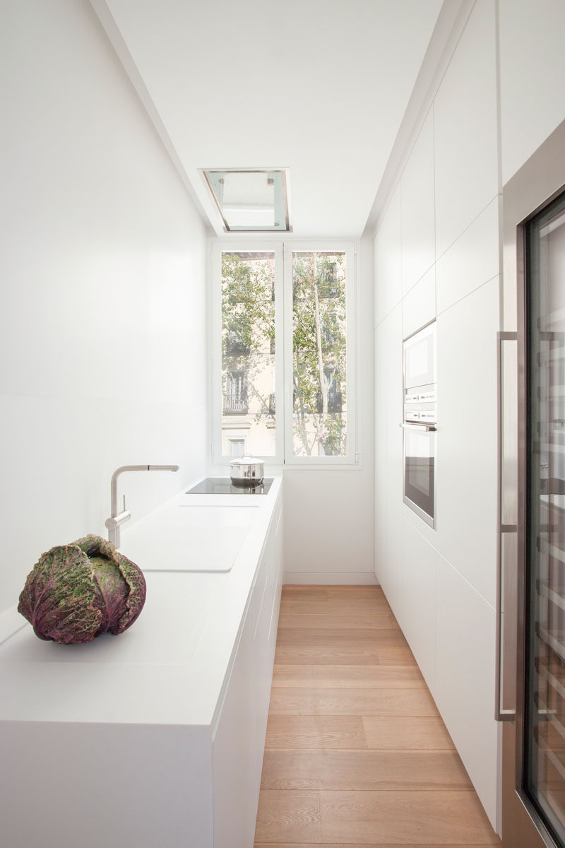 This galley kitchen has minimalist white cabinets on one side for storage, while the opposite side of the modern kitchen has the countertop, sink and cooktop. As the kitchen is white, the light from the tall windows at the end of the kitchen reflects throughout the space, making it bright and airy.