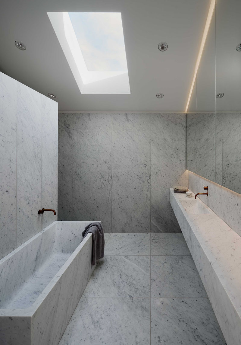 The design of this modern bathroom includes plenty of Carrara marble, with the material being used for the walls, floors, bathtub and vanity. A skylight lets natural light into the space, while a backlit mirror provides additional lighting when it gets dark outside.
