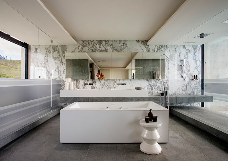 This modern master bathroom has an open-plan layout. In the middle of the bathroom is a freestanding bathtub, while the vanity extends into the large, glass enclosed shower that enjoys views of the city.