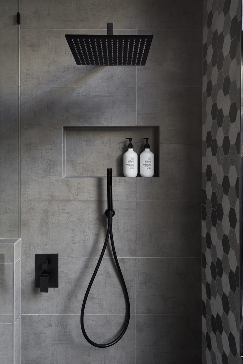 In this modern bathroom, the shower has a matte black rainfall shower head and a hand held shower head, as well as a tiled built-in shelf.