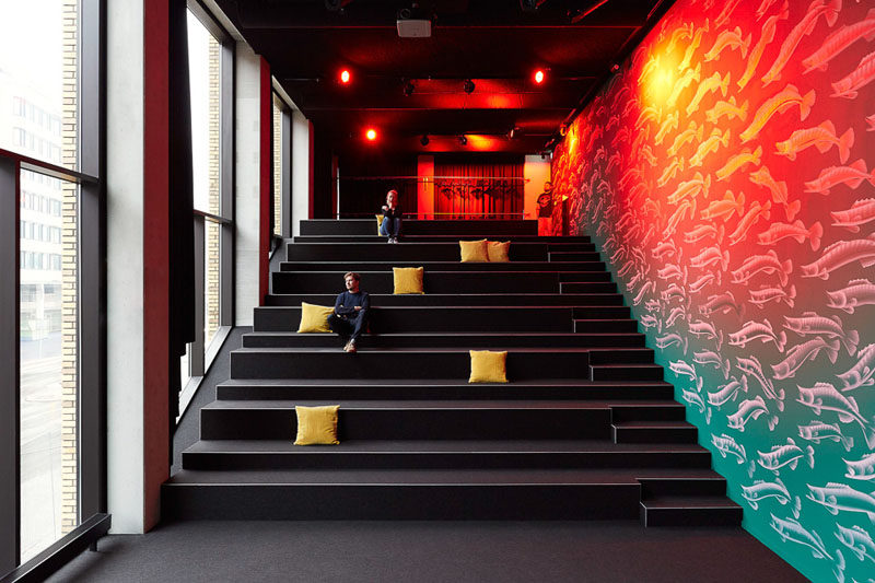 In this modern hotel design, there's an auditorium with rows of tiered seating for talks and lectures to be delivered in an area with lots of natural light and a unique colorful mural along one wall.