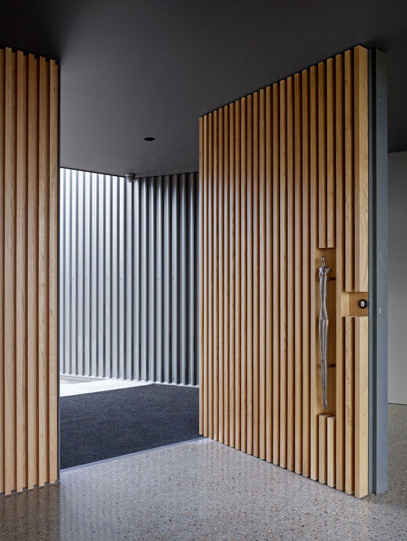 This large pivoting front door covered in light wood slats has a sculptural door handle set into the wood to welcome you into the home.