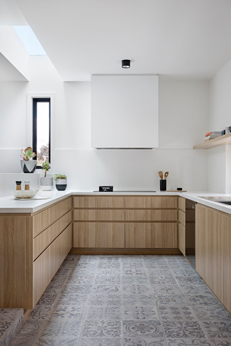In this modern kitchen, there's a skylight above the white counters, and light wood cabinets help to keep the space bright. A light grey patterned floor tile adds some interest, while the exhaust fan above the oven is white so that it blends in with the wall.