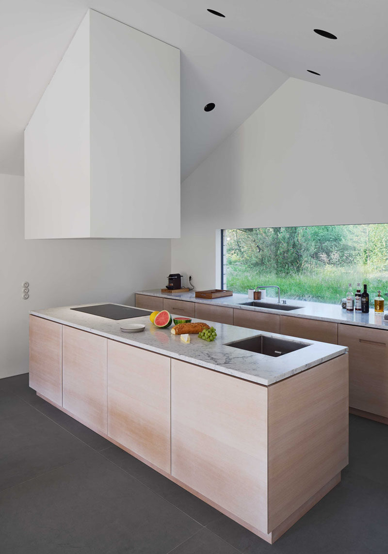 In this modern light wood and stone kitchen, there are high ceilings making the space feel lofty and open. A large window allows for views of the outdoors and natural light to flood the countertop.