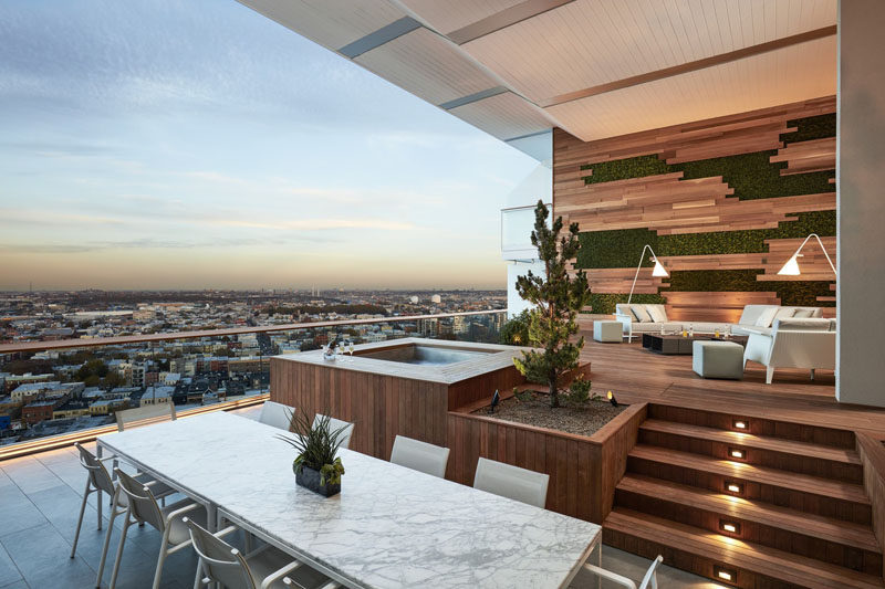 This large modern furnished balcony features plenty of natural wood and includes an elevated seating area with a feature wall made from wood strips and patches of plants, a white and grey outdoor lounge with floor lamps, an open air Jacuzzi surrounded by hardwood and a long white outdoor dining table.