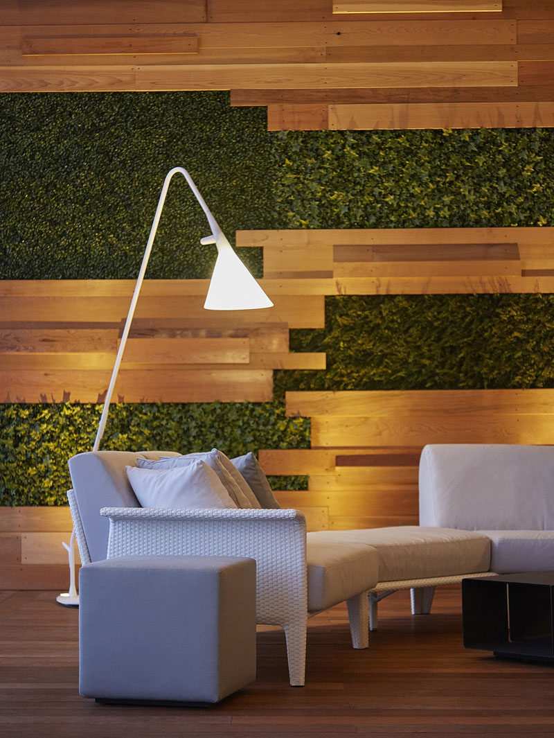 This large modern furnished balcony features an elevated seating area with a feature wall made from wood strips and patches of plants as well as an white and grey outdoor lounge with floor lamps.