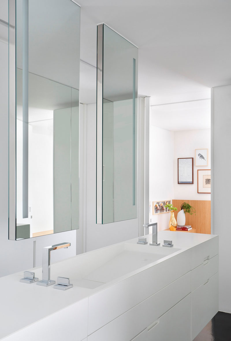 This modern ensuite bathroom that also doubles as a dressing room, features white Corian countertops and double vanity with two hanging mirrors that help brighten the space by reflecting back the all white cabinetry.