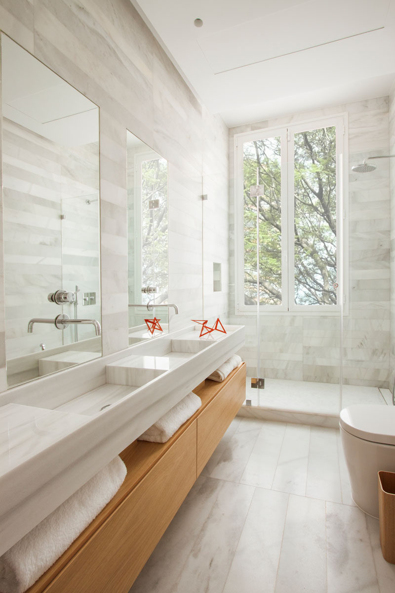 In this modern bathroom there's a wooden bathroom vanity with open shelving, that has double sinks with tall rectangular mirrors above each one. A glass shower surround allows the light from the vertical windows to pass through and fill the room, while stone tile adds a soft natural touch to the space.