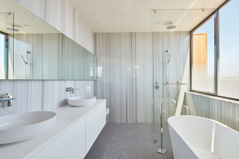 This modern bathroom has grey stone walls, a floating white double vanity, a rainfall shower with glass surround and a standalone bathtub.