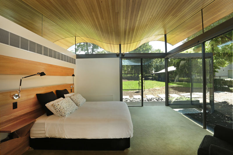 In this modern bedroom there's a softly rolling ceiling and large floor-to-ceiling windows and sliding glass doors that leads out into the backyard. The wood headboard behind the beds provides built-in side tables, lights, and a shelf that can be used to display anything from photos to flowers to art.