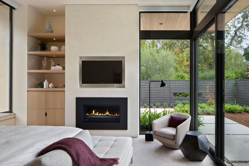 In this modern bedroom, there's a built-in fireplace with a recessed TV positioned above it, and large floor-to-ceiling windows keep the bedroom cozy and bright. Built-in shelving and storage also makes sure that the room stays organized and tidy while the built-in lights in the shelves highlight personal decorative items.