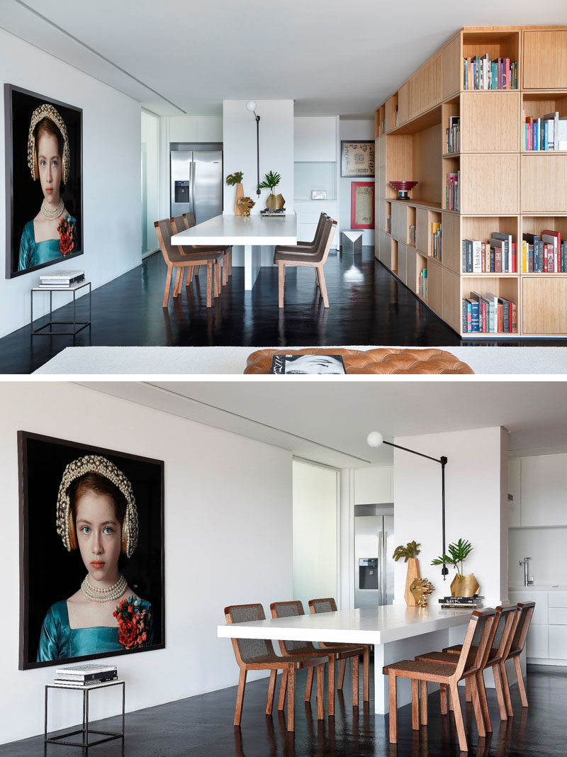 In this modern kitchen and dining area, a long white dining table is attached to a central wall, and the kitchen at the back of the dining room features all white cabinetry and stainless steel appliances.