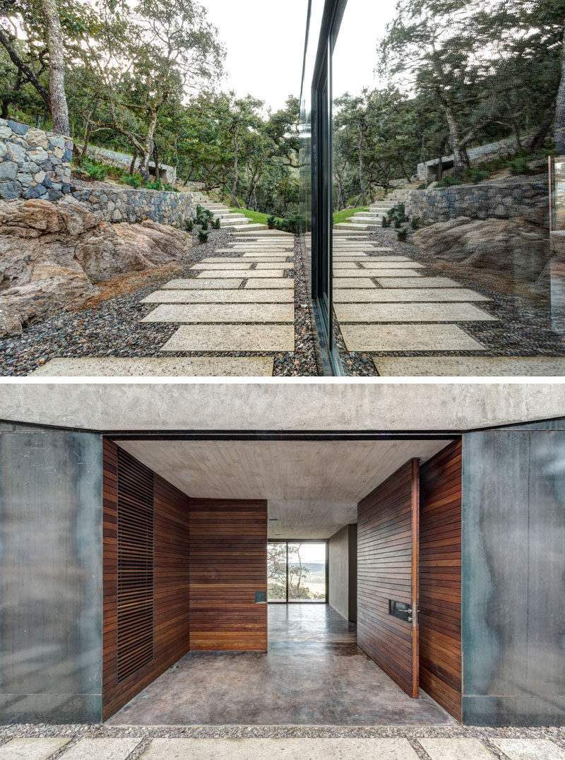 This modern house has a stone path that leads from the garage to the front entryway. The entryway, including the front door, are covered in wood, breaking up the harshness of the surrounding steel and the concrete overhead.