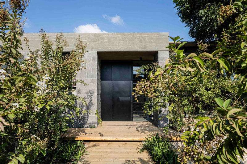 This modern house has a light wood path leading to a black steel front door, that's surrounded by concrete blocks and a concrete roof.