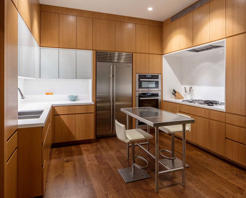 In this U-shaped modern kitchen, wood cabinets are broken up by white countertops, tiles and additional cabinets. A small stainless steel island ties in with the stainless steel appliances.