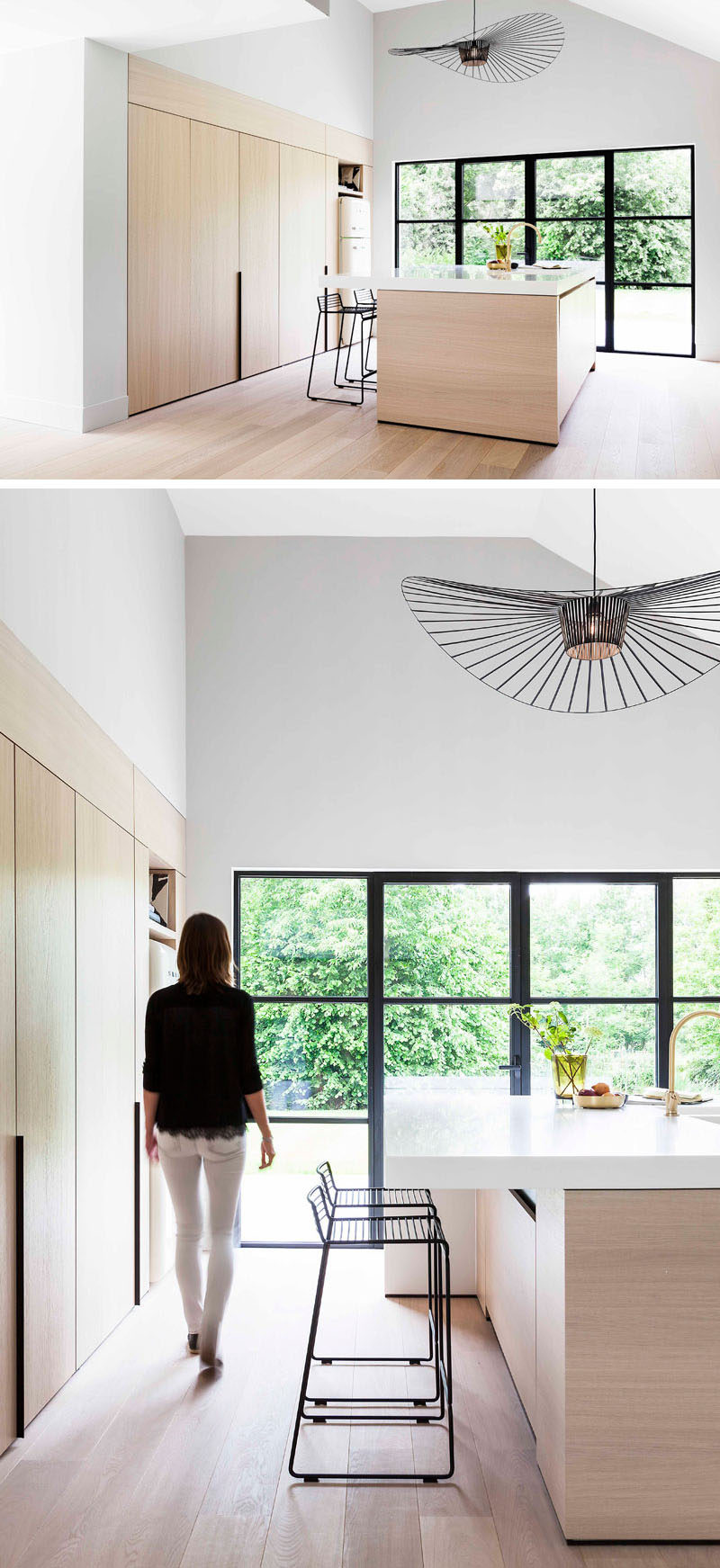 This modern and minimalist light wood and white kitchen features black accents like stools and window frames, a large central island, foldaway cabinet doors, and a pantry with hidden lighting.