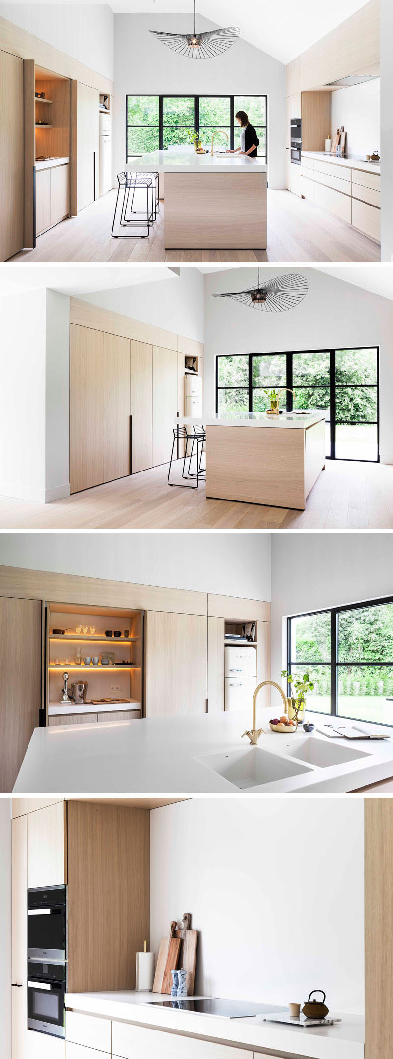 This modern and minimalist light wood and white kitchen features black accents like stools and window frames, a large central island, foldaway cabinet doors, and a pantry with hidden lighting.