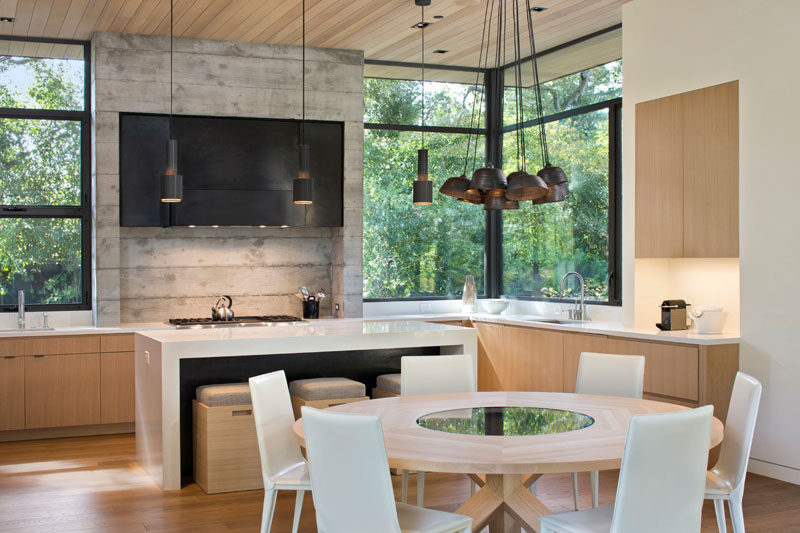 In this modern kitchen, the concrete and metal stove surround, wood cabinets, and white island and countertops create a modern look that's kept natural and bright thanks to the large windows looking out into the trees, and the pendant lights hanging above the island and dining table.