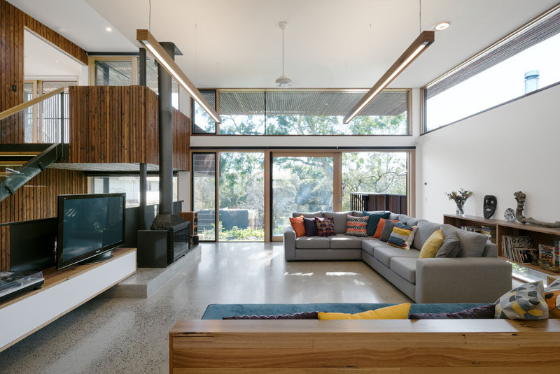 In the living room of this modern house, large windows look out onto the backyard and fill the room with natural light while the wood along the one side creates a cozy, warm, and natural feel in the space.